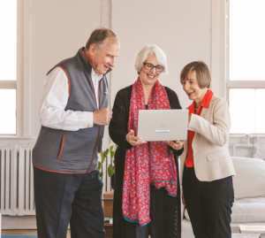 Three older adults smiling as they look at a laptop together in a bright office.