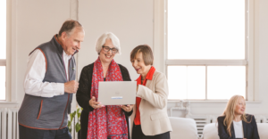 Three older adults smiling as they look at a laptop together in a bright office.