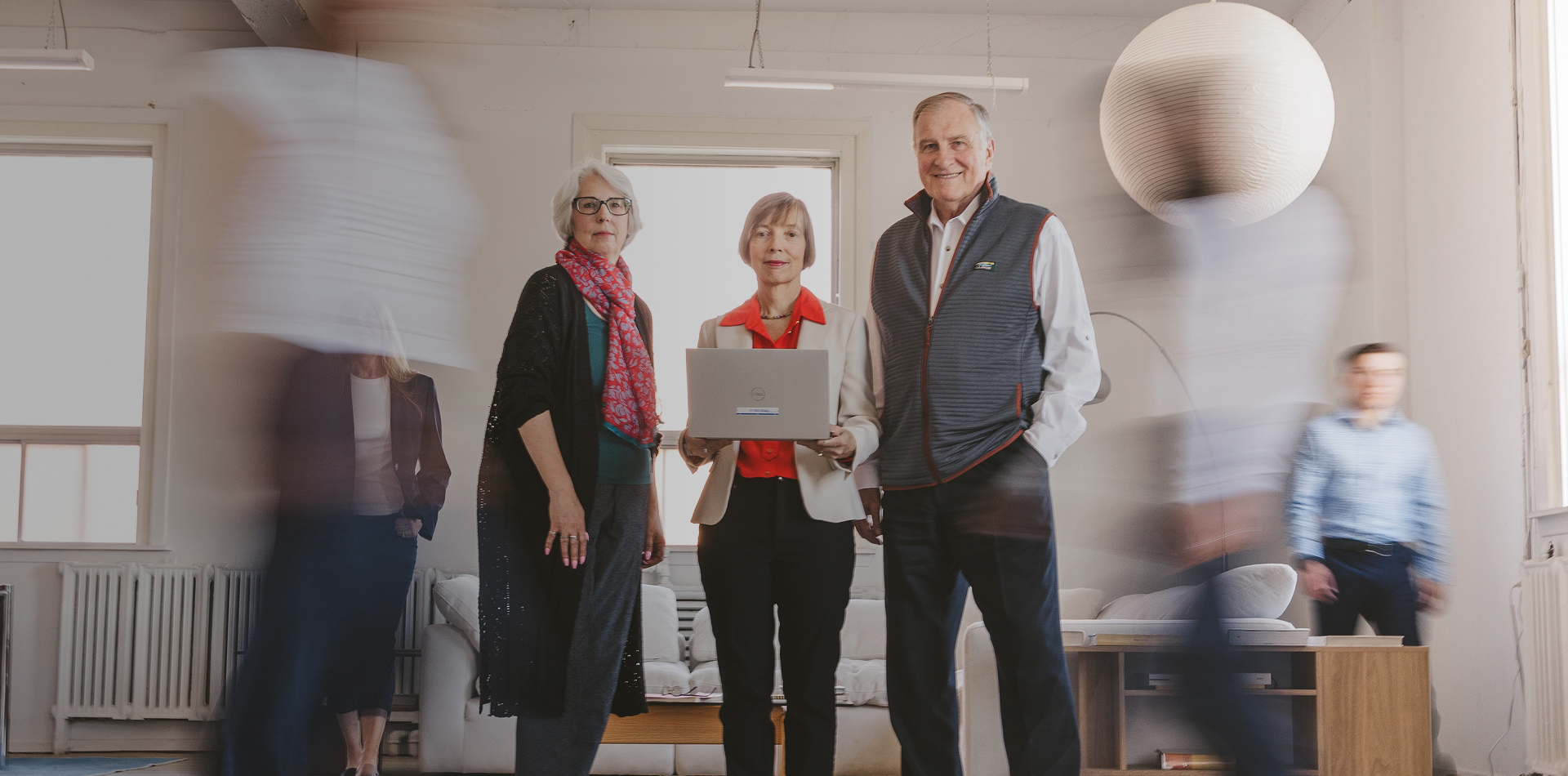 Three adults posing with a laptop as coworkers move through a office.