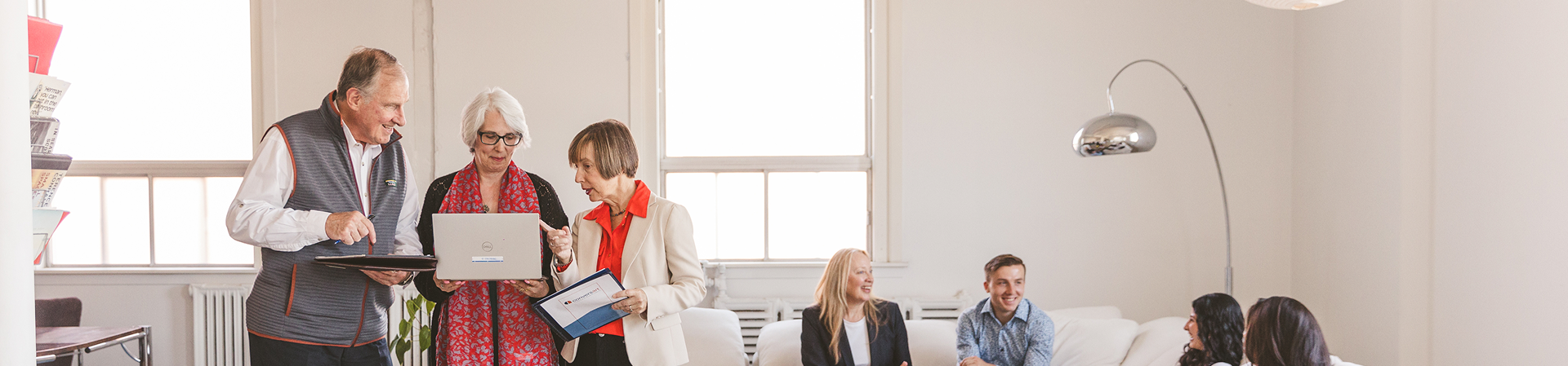 Three older adults looking at a laptop while colleagues talk in a bright office.