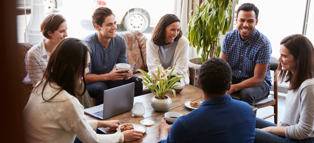 Seven coworkers smiling and talking around a table with a laptop and coffee.